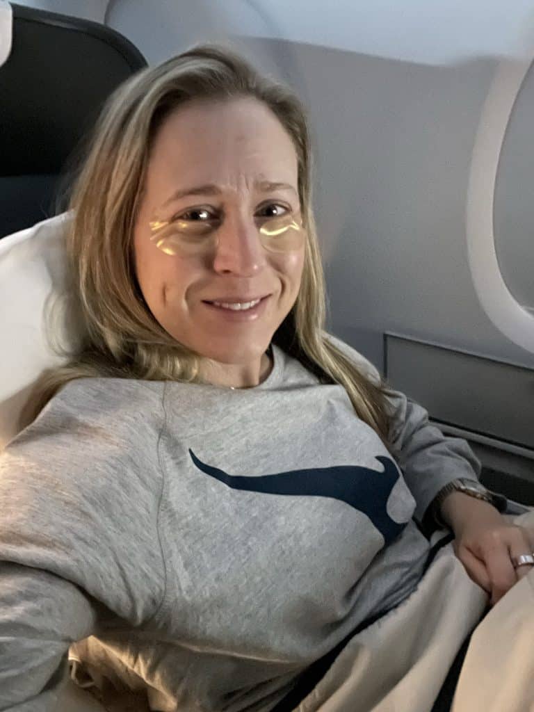 A person with gold eye patches is smiling while lying back in an airplane seat. They are wearing a grey sweatshirt with a blue logo and are covered with a light-colored blanket.