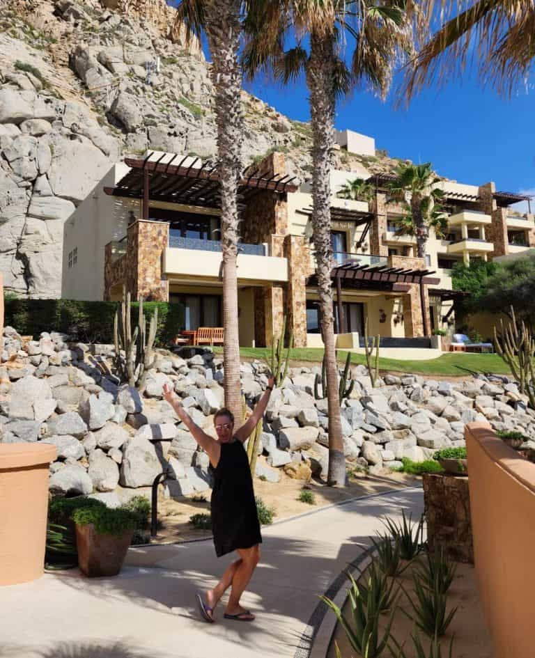 A person in a black dress joyfully poses with raised arms on a pathway lined with cacti, in front of a hillside house with stone accents and terraces. Palm trees and blue sky enhance the tropical setting.