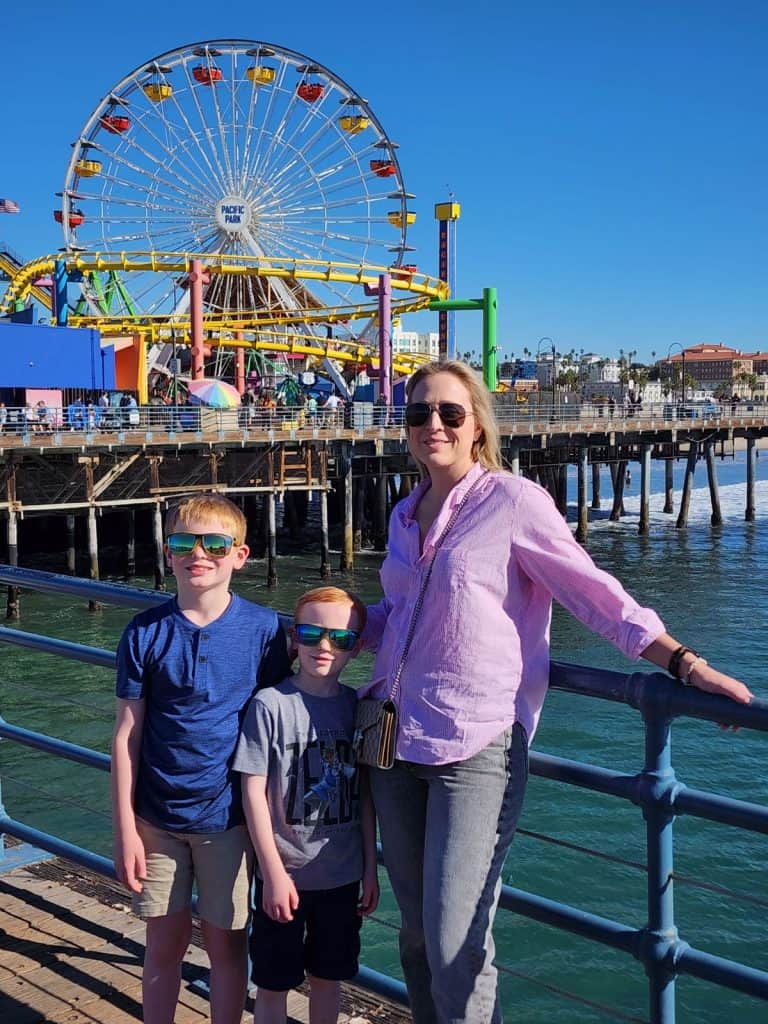 A woman and two children stand on a pier with a colorful Ferris wheel in the background under a clear blue sky. The woman is wearing sunglasses and a pink shirt, while the children are in casual shirts and shorts, also wearing sunglasses.