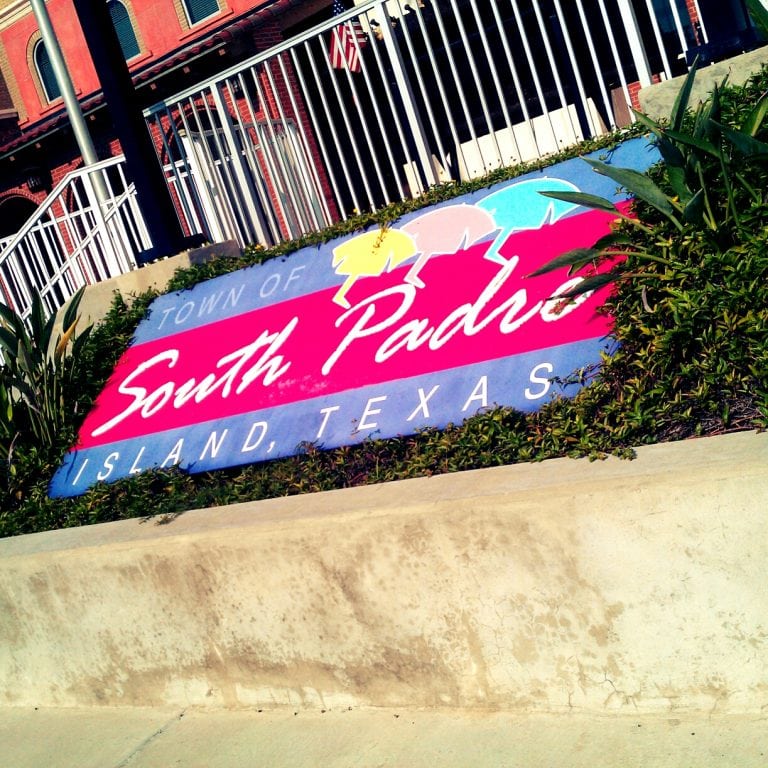 A welcome sign for the town of South Padre Island, Texas, featuring a bold pink and blue background with "South Padre" written in white cursive and "Island, Texas" in block letters. The sign is partially surrounded by green plants and includes illustrations of three beach umbrellas. A white metal fence and a red brick building are visible in the background.